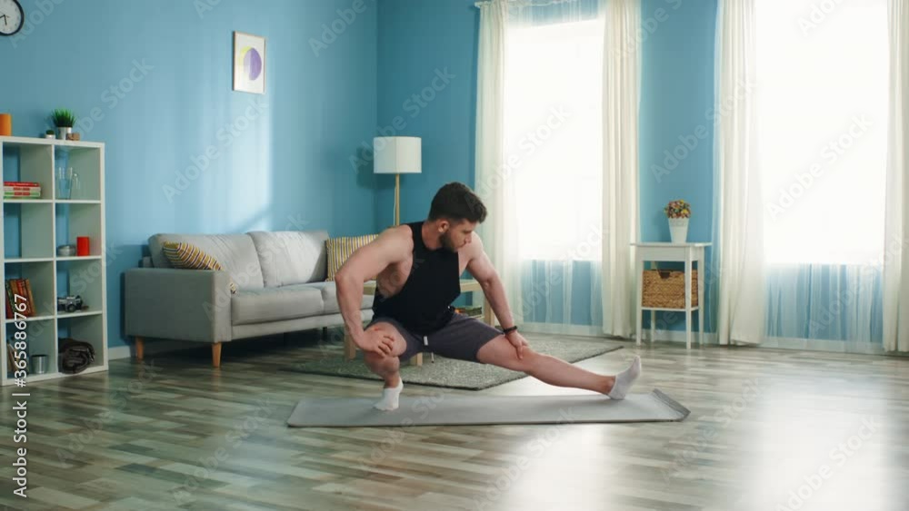 Strong athletic man is doing morning exercise, warming up in living room, stretching his legs, getting ready to daily workout, putting efforts to maintain perfect body shape, Slow motion. Strong athletic man is doing morning exercise, warming up in living room, stretching his legs, getting ready to daily workout, putting efforts to maintain perfect body shape, Slow motion.