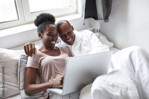 beautiful happy african american couple using laptop on bed, the look at screen of laptop and talk. morning