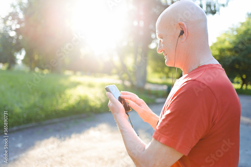 Smiling old man in casual wear and white headphones listening music from his smartphone in the park.