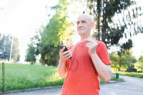 Smiling old man in casual wear and white headphones listening music from his smartphone in the park.