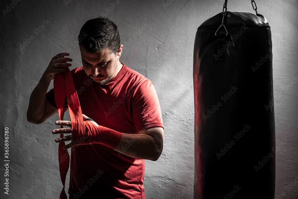 Boxeador joven principiante con camiseta roja poniendose cinta en las manos  junto a un saco negro