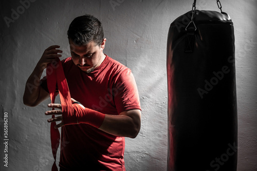 Boxeador joven principiante con camiseta roja poniendose cinta en las manos junto a un saco negro en su garaje con guantes de boxeo negros, iluminado con luz intensa de flash y fondo negro photo