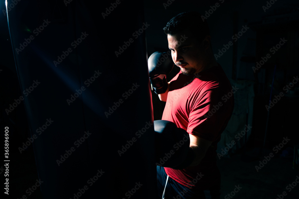 Boxeador joven principiante con camiseta roja golpeando un saco negro en su garaje con guantes de boxeo negros, iluminado con luz intensa de flash y fondo negro Boxeador joven principiante con camiseta roja golpeando un saco negro en su garaje con guantes de boxeo negros, iluminado con luz intensa de flash y fondo negro