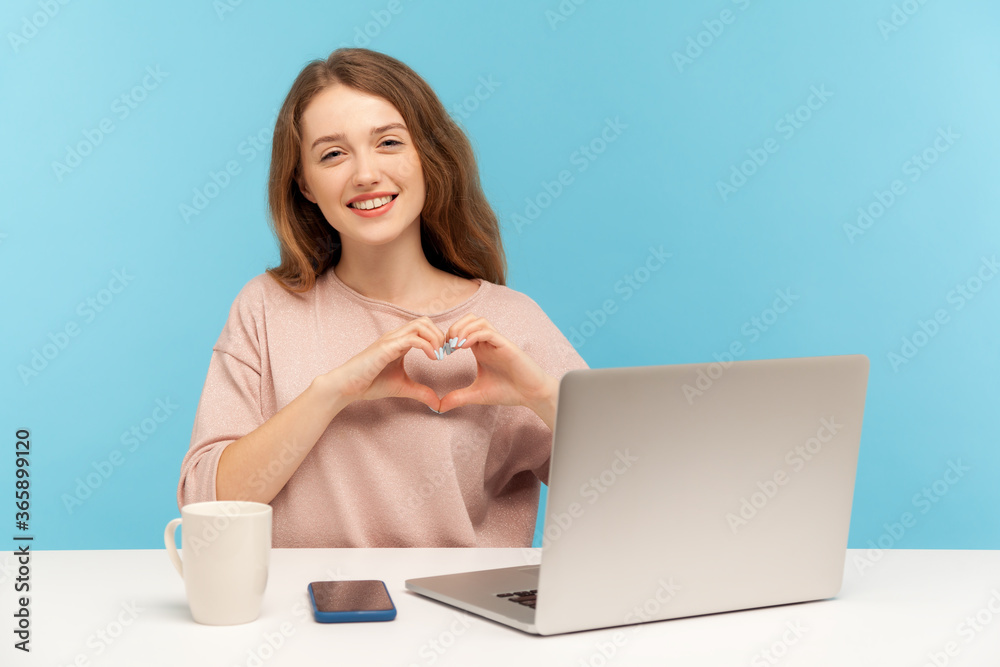 Feminine young pretty woman sitting at workplace with laptop and making heart shape with hands, smiling lovely, expressing romantic amorous feelings. indoor studio shot isolated on blue background Feminine young pretty woman sitting at workplace with laptop and making heart shape with hands, smiling lovely, expressing romantic amorous feelings. indoor studio shot isolated on blue background