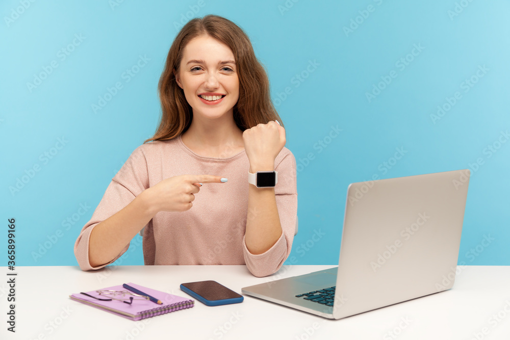 Look at clock, time to work! Positive businesswoman sitting workplace and pointing wrist watch, looking at camera with toothy smile, reminding late hour. indoor studio shot isolated on blue background Look at clock, time to work! Positive businesswoman sitting workplace and pointing wrist watch, looking at camera with toothy smile, reminding late hour. indoor studio shot isolated on blue background