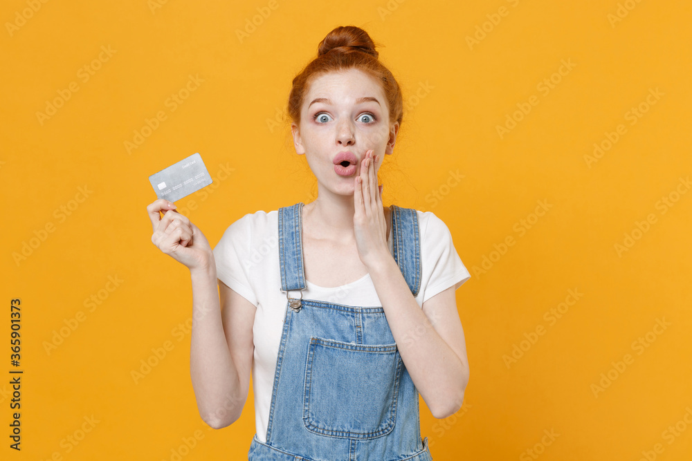 Amazed young readhead girl in casual denim clothes white t-shirt isolated on yellow background studio portrait. People lifestyle concept. Mock up copy space. Hold credit bank card, put hand on cheek. Amazed young readhead girl in casual denim clothes white t-shirt isolated on yellow background studio portrait. People lifestyle concept. Mock up copy space. Hold credit bank card, put hand on cheek.
