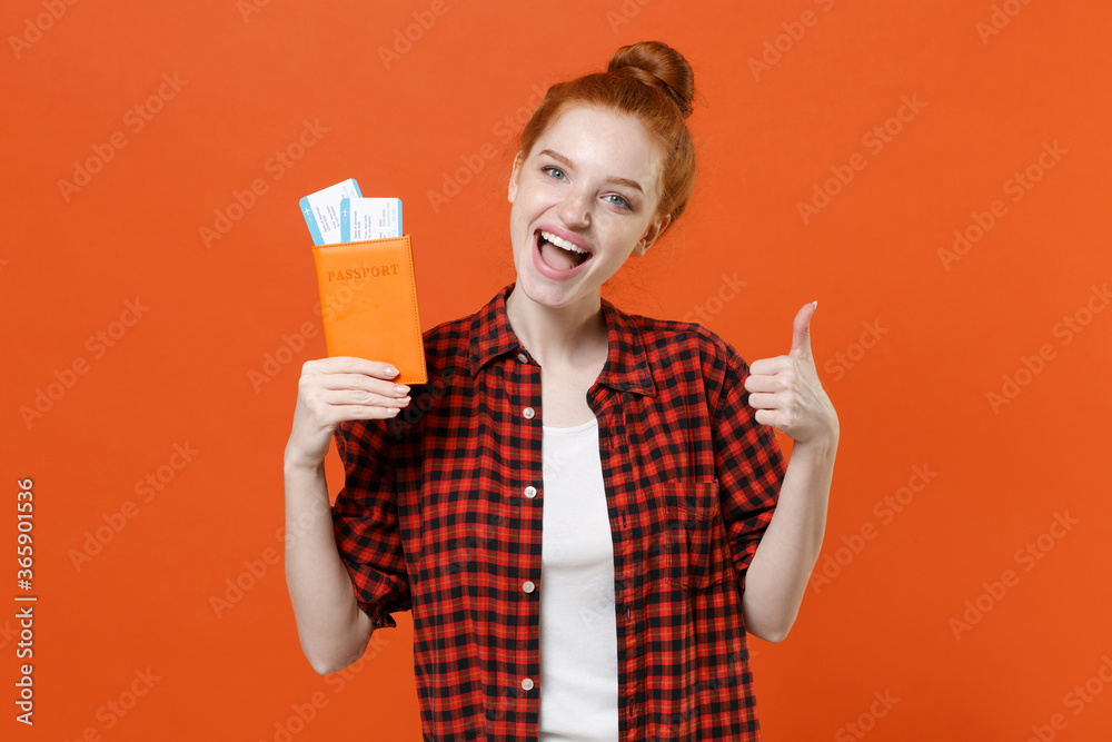 Excited young girl in red checkered shirt isolated on orange background. Passenger traveling abroad to travel on weekends getaway. Air flight journey concept. Hold passport tickets showing thumb up. Excited young girl in red checkered shirt isolated on orange background. Passenger traveling abroad to travel on weekends getaway. Air flight journey concept. Hold passport tickets showing thumb up.