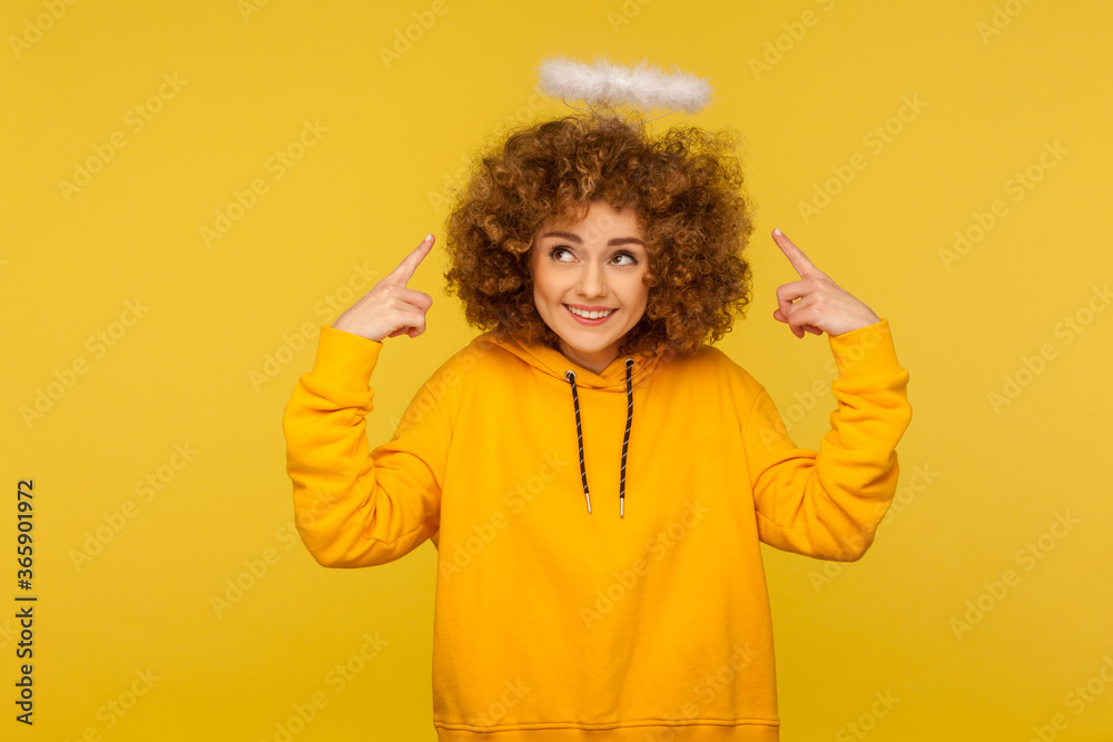 Portrait of charming angel with halo, kind curly-haired hipster woman pointing at saint nimbus over her head and smiling with obedient modest expression. studio shot isolated on yellow background Portrait of charming angel with halo, kind curly-haired hipster woman pointing at saint nimbus over her head and smiling with obedient modest expression. studio shot isolated on yellow background