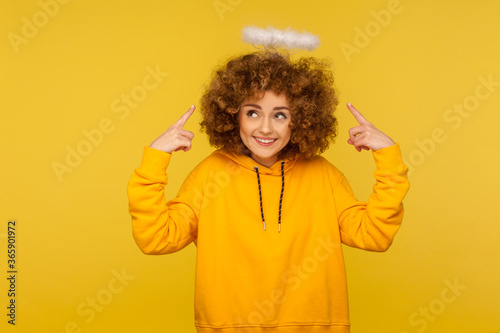 Portrait of charming angel with halo  kind curly-haired hipster woman pointing at saint nimbus over her head and smiling with obedient modest expression. studio shot isolated on yellow background