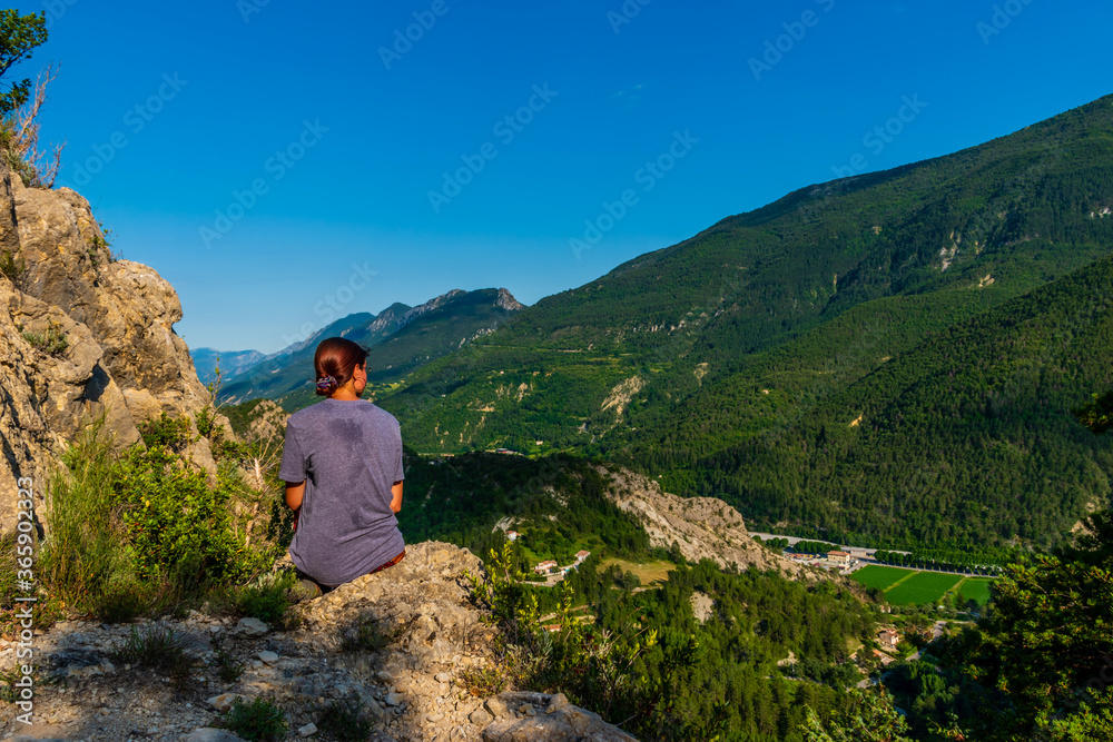 A full body shot of a thoughtful unrecognizable young Caucasian redhead hiker sitting on a boulder on a hiking path and enjoying the view of the French Alps during sunset