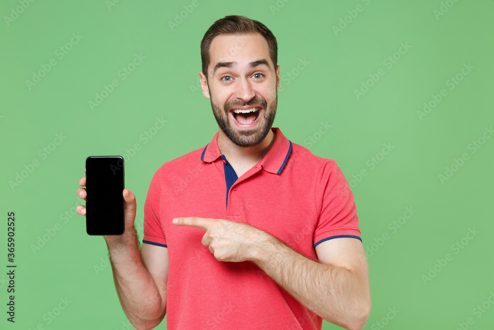 Excited young bearded man guy in casual red pink t-shirt posing isolated on green background. People lifestyle concept. Mock up copy space. Point index finger on mobile phone with blank empty screen. Excited young bearded man guy in casual red pink t-shirt posing isolated on green background. People lifestyle concept. Mock up copy space. Point index finger on mobile phone with blank empty screen.