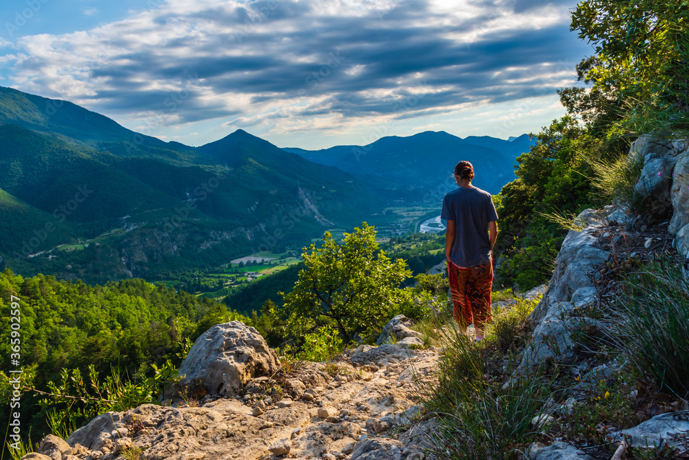 Naklejka premium A full body shot of a thoughtful unrecognizable young Caucasian redhead hiker standing on a hiking path and enjoying the view of the French Alps during sunset