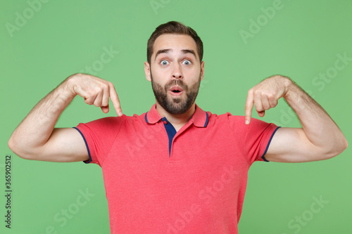 Shocked young bearded man guy in casual red pink t-shirt posing isolated on green background studio portrait. People sincere emotions lifestyle concept. Mock up copy space. Point index fingers down.