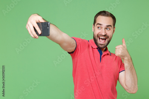 Excited young bearded man guy in casual red pink t-shirt posing isolated on green background studio. People lifestyle concept. Mock up copy space. Doing selfie shot on mobile phone, showing thumb up.