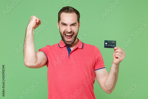 Overjoyed young bearded man guy in casual red pink t-shirt posing isolated on green background studio portrait. People lifestyle concept. Mock up copy space Hold credit bank card doing winner gesture.