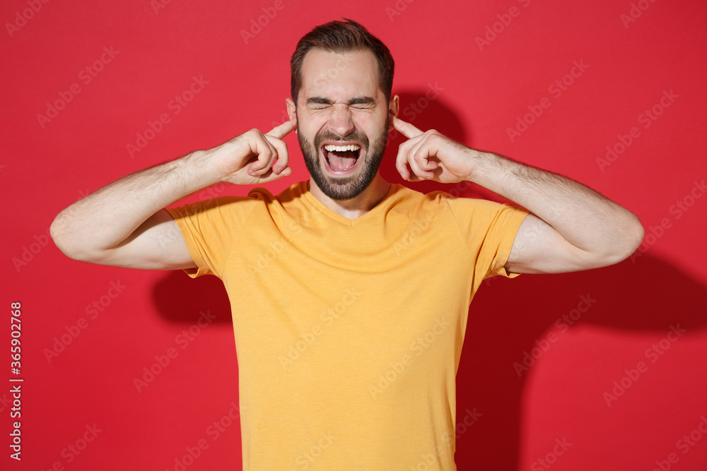 Crazy young bearded man guy in casual yellow t-shirt posing isolated on red wall background. People lifestyle concept. Mock up copy space. Covering ears with fingers, keeping eyes closed, screaming. Crazy young bearded man guy in casual yellow t-shirt posing isolated on red wall background. People lifestyle concept. Mock up copy space. Covering ears with fingers, keeping eyes closed, screaming.
