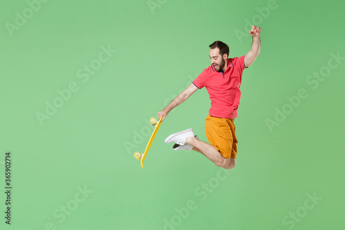 Funny young bearded man guy in casual red pink t-shirt posing isolated on green wall background studio portrait. People emotions lifestyle concept. Mock up copy space. Jumping, hold yellow skateboard. © ViDi Studio