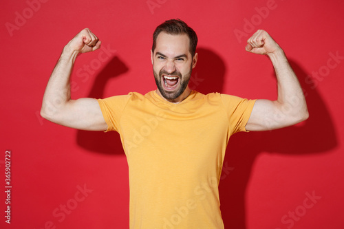 Strong funny young bearded man guy in casual yellow t-shirt posing isolated on red background studio portrait. People sincere emotions lifestyle concept. Mock up copy space. Showing biceps, muscles.