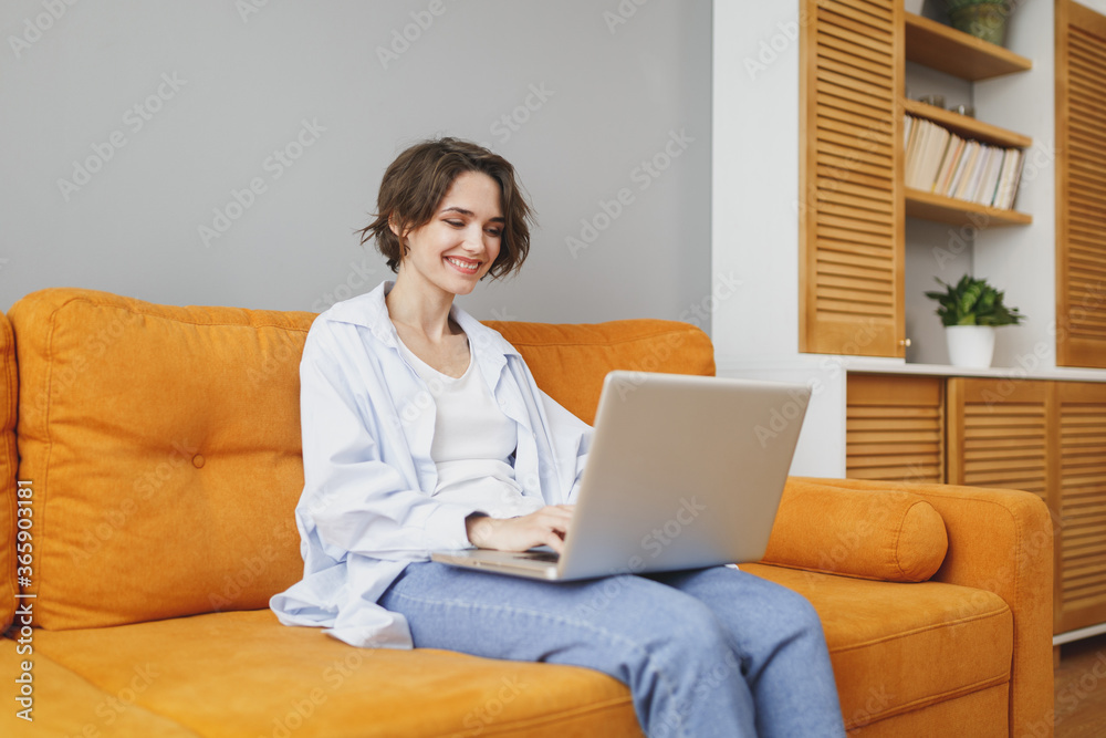 Smiling young woman girl in casual clothes sitting on couch spending time in living room at home. Rest relax good mood leisure lifestyle concept. Mock up copy space. Working on laptop pc computer. Smiling young woman girl in casual clothes sitting on couch spending time in living room at home. Rest relax good mood leisure lifestyle concept. Mock up copy space. Working on laptop pc computer.