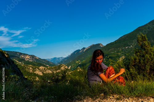 A full body shot of a curious young Caucasian redhead hiker sitting on the ground looking at a plant in the French Alps during sunset