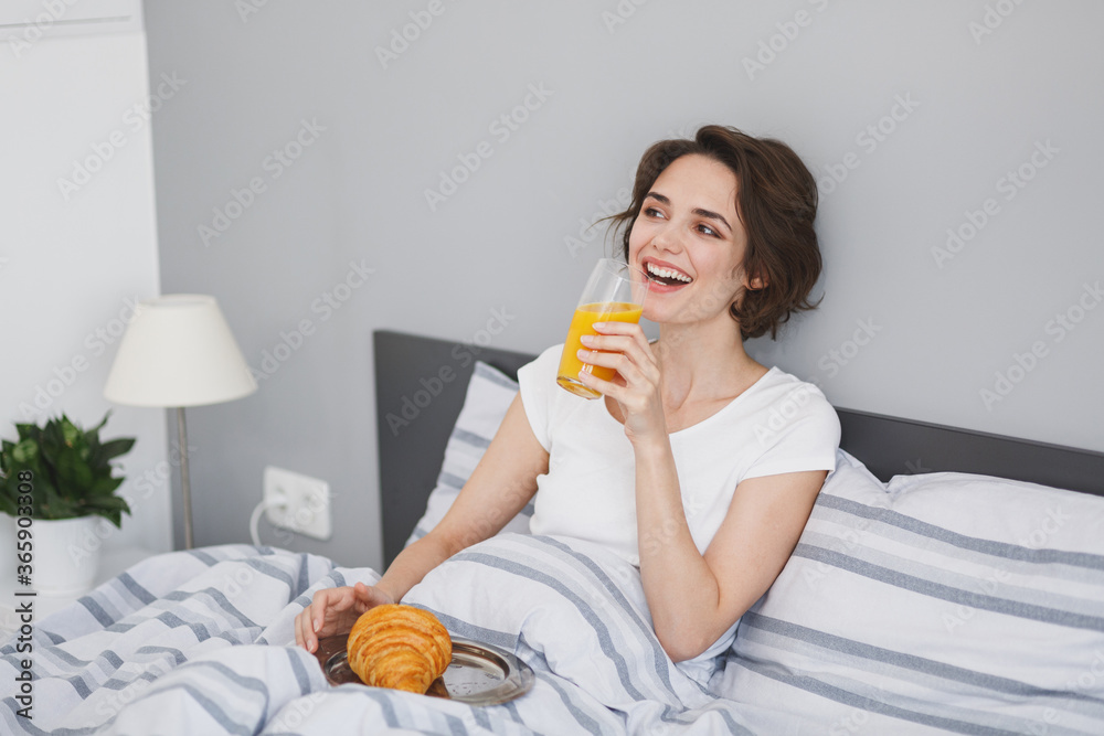 Cheerful young brunette woman girl in white t-shirt lying in bed spending time in bedroom at home. Rest relax good mood lifestyle concept. Having breakfast, hold plate with croissant, glass of juice. Cheerful young brunette woman girl in white t-shirt lying in bed spending time in bedroom at home. Rest relax good mood lifestyle concept. Having breakfast, hold plate with croissant, glass of juice.