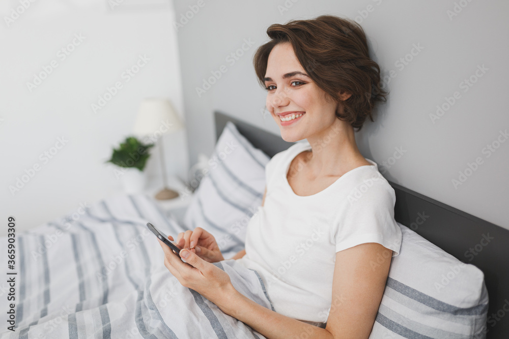 Beautiful young woman in white t-shirt lying in bed with striped sheet pillow blanket spending time in bedroom at home. Rest relax good mood lifestyle concept. Using mobile phone, typing sms message. Beautiful young woman in white t-shirt lying in bed with striped sheet pillow blanket spending time in bedroom at home. Rest relax good mood lifestyle concept. Using mobile phone, typing sms message.