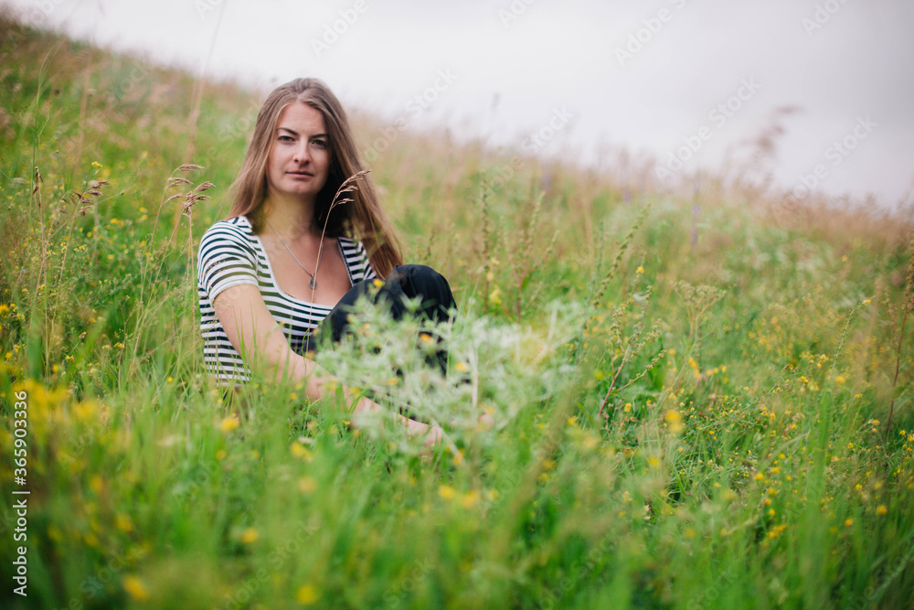 Beautiful girl in a black long skirt and striped T-shirt sitting on the slope of a green meadow Beautiful girl in a black long skirt and striped T-shirt sitting on the slope of a green meadow