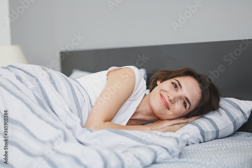 Beautiful smiling young woman in white t-shirt sleeping lying in bed with striped sheet pillow blanket spending time in bedroom at home. Rest relax good mood lifestyle concept. Mock up copy space.