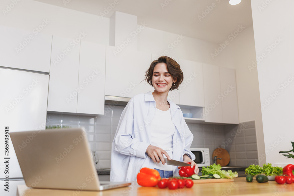 Smiling young housewife woman girl in white casual clothes using laptop computer looking at recipe preparing vegetable salad cooking food in light kitchen at home. Dieting healthy lifestyle concept.