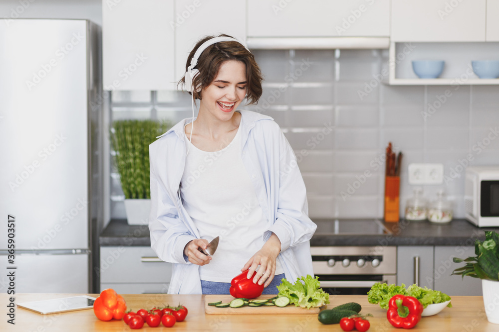 Cheerful young housewife woman in casual clothes preparing vegetable salad cooking food in light kitchen at home. Dieting healthy lifestyle concept. Mock up copy space. Listen music with headphones.