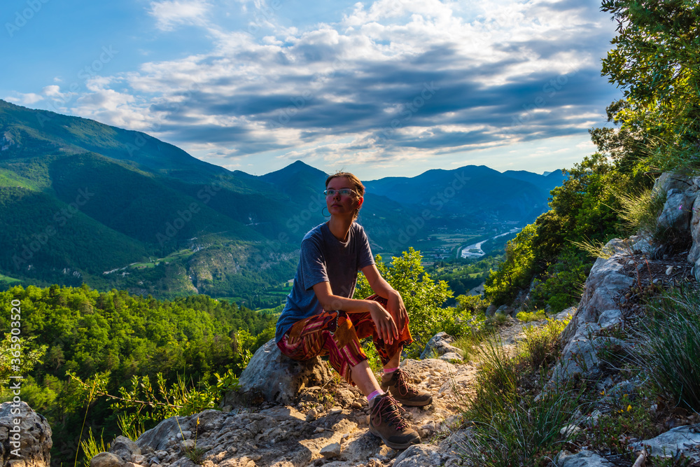 Naklejka premium A full body shot of a thoughtful young Caucasian redhead hiker sitting on a boulder on a hiking path and enjoying the view of the French Alps during sunset
