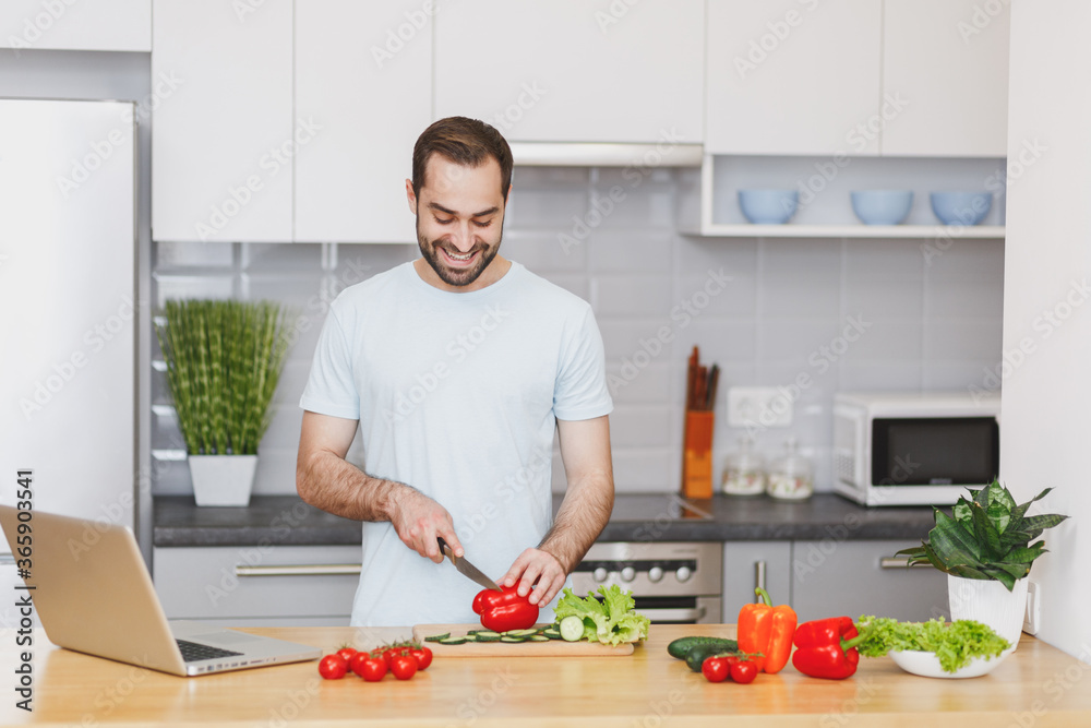 Smiling handsome bearded young man guy in white casual t-shirt posing preparing vegetable salad cooking food in light kitchen at home. Dieting healthy lifestyle concept. Mock up copy space. Smiling handsome bearded young man guy in white casual t-shirt posing preparing vegetable salad cooking food in light kitchen at home. Dieting healthy lifestyle concept. Mock up copy space.
