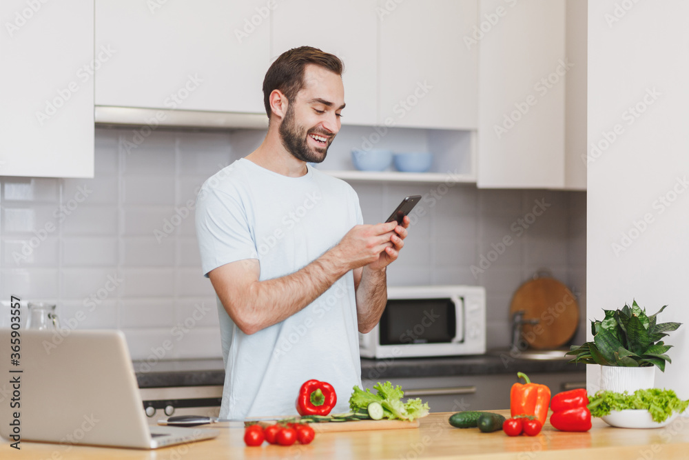 Smiling young bearded man in white t-shirt using mobile phone looking at recipe preparing vegetable salad cooking food in light kitchen at home. Dieting healthy lifestyle concept. Mock up copy space. Smiling young bearded man in white t-shirt using mobile phone looking at recipe preparing vegetable salad cooking food in light kitchen at home. Dieting healthy lifestyle concept. Mock up copy space.