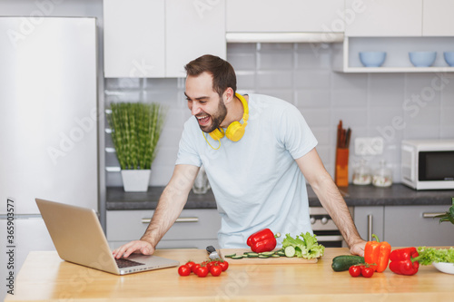 Funny young bearded man in casual t-shirt with headphones using laptop computer looking at recipe preparing vegetable salad cooking food in light kitchen at home. Dieting healthy lifestyle concept.