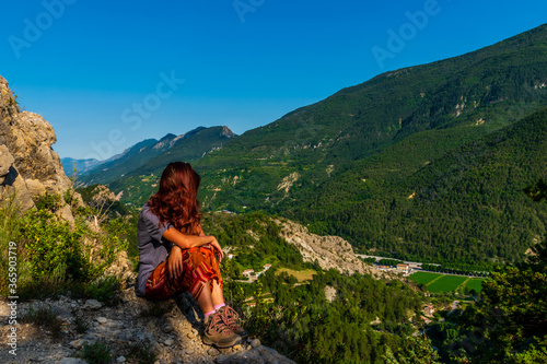 A full body shot of a thoughtful unrecognizable young Caucasian redhead hiker sitting on a boulder on a hiking path and enjoying the view of the French Alps during sunset © k.dei