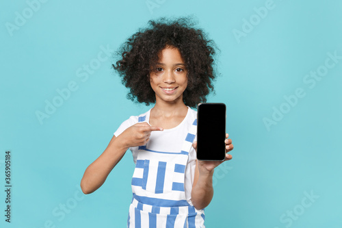 Smiling little african american kid girl 12-13 years old in striped clothes isolated on blue background. Childhood lifestyle concept. Pointing index finger on mobile phone with blank empty screen.