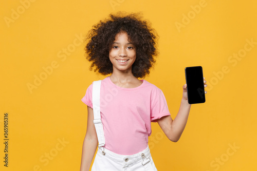 Smiling little african american kid girl 12-13 years old in pink t-shirt isolated on yellow wall background studio. Childhood lifestyleconcept. Mock up copy space. Hold mobile phone with blank screen. photo