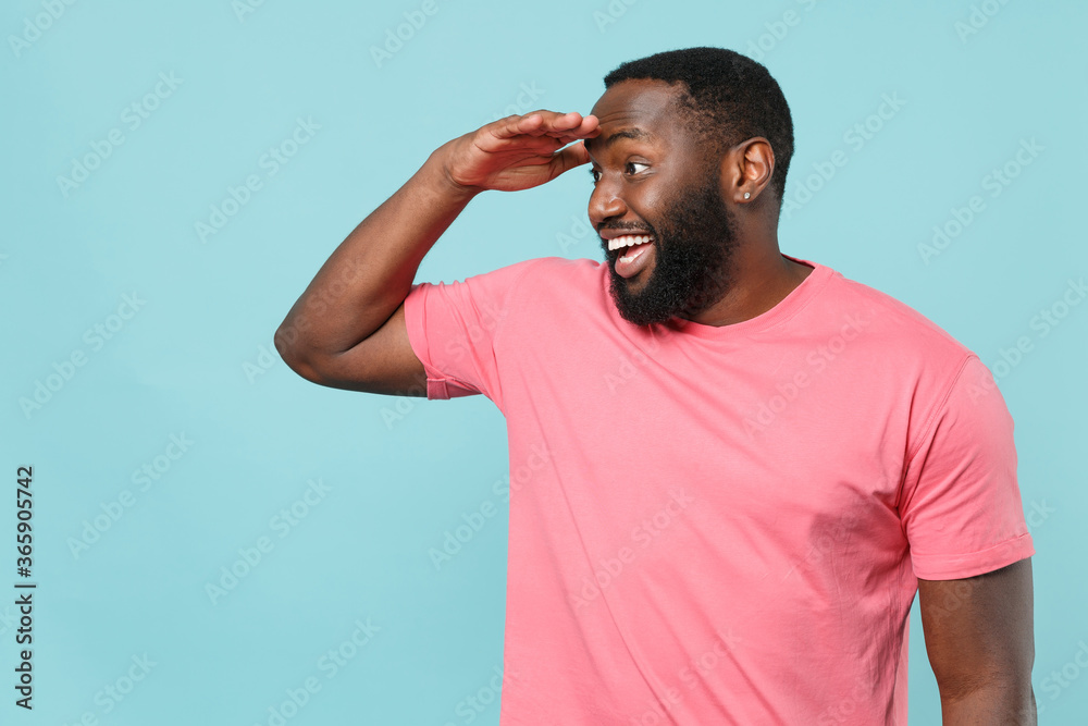 Excited young african american man guy in casual pink t-shirt posing isolated on blue background studio. People lifestyle concept. Mock up copy space. Hold hand at forehead looking far away distance. Excited young african american man guy in casual pink t-shirt posing isolated on blue background studio. People lifestyle concept. Mock up copy space. Hold hand at forehead looking far away distance.
