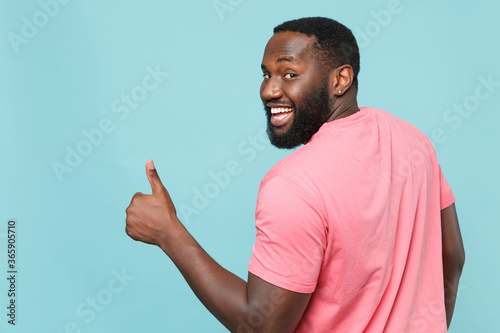 Back rear view of funny young african american man guy in casual pink t-shirt posing isolated on blue background studio. People lifestyle concept. Mock up copy space. Showing thumb up, looking camera.