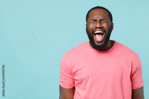 Frustrated young african american man guy in casual pink t-shirt isolated on blue wall background studio portrait. People emotions lifestyle concept. Mock up copy space. Keeping eyes closed screaming.