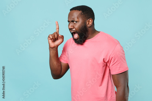 Angry young african american man guy in casual pink t-shirt isolated on blue wall background studio portrait. People lifestyle concept. Mock up copy space. Pointing index finger up screaming swearing.