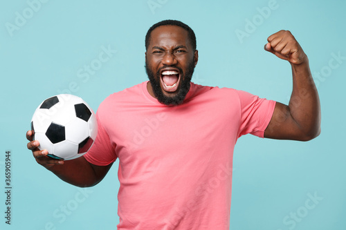 Screaming african american man guy football fan in pink t-shirt isolated on blue background. Sport family leisure lifestyle concept. Cheer up support favorite team with soccer ball, clenching fist.