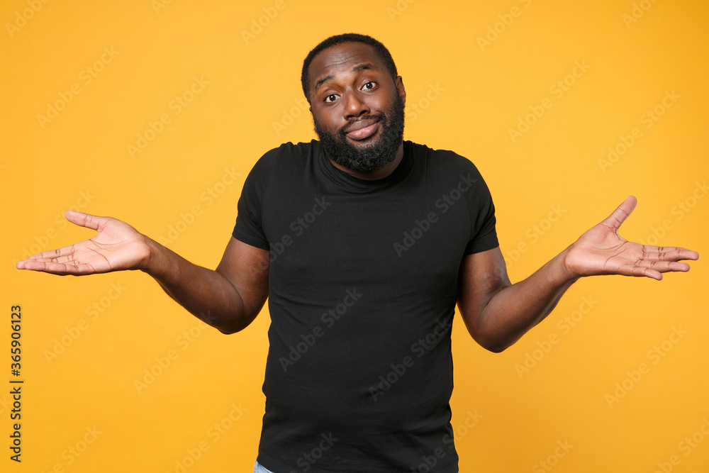 Confused african american man football fan in casual black t-shirt isolated on yellow wall background studio portrait. People sincere emotions lifestyle concept. Mock up copy space. Spreading hands. Confused african american man football fan in casual black t-shirt isolated on yellow wall background studio portrait. People sincere emotions lifestyle concept. Mock up copy space. Spreading hands.