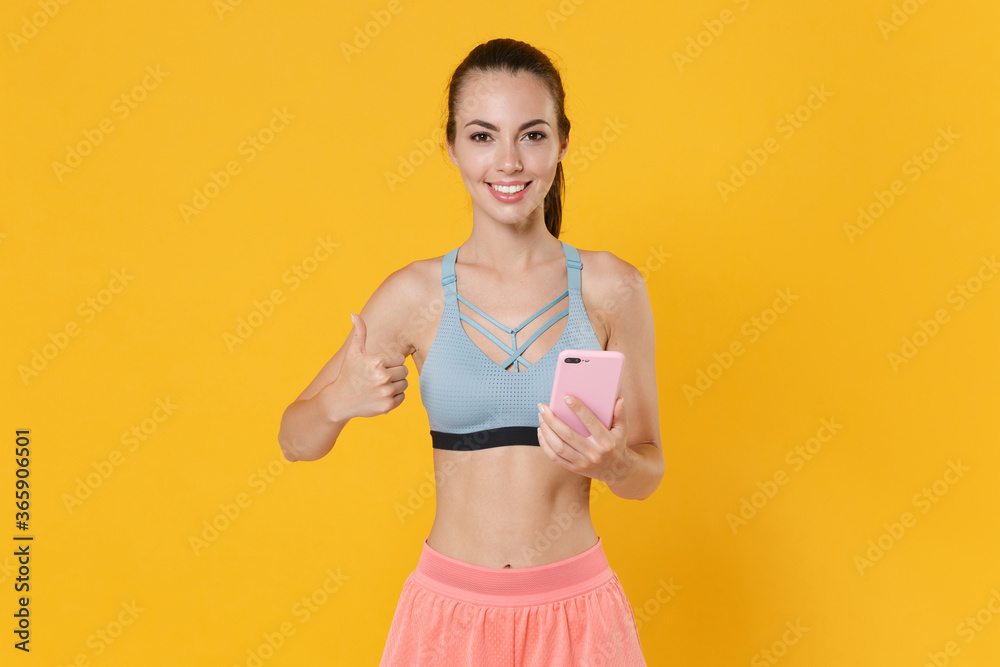 Smiling young fitness sporty woman girl in sportswear working out isolated on yellow background. Workout sport motivation lifestyle concept. Mock up copy space. Using mobile phone, showing thumb up. Smiling young fitness sporty woman girl in sportswear working out isolated on yellow background. Workout sport motivation lifestyle concept. Mock up copy space. Using mobile phone, showing thumb up.