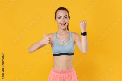 Smiling young fitness sporty girl in sportswear working out isolated on yellow background. Workout sport motivation lifestyle concept. Mock up copy space. Wearing smart watch on hand showing thumb up.