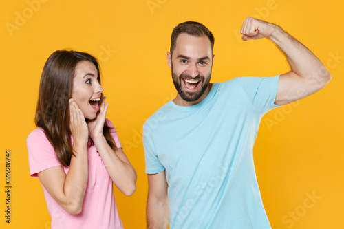 Excited young couple friends guy girl in blue pink t-shirts isolated on yellow background studio portrait. People lifestyle concept. Mock up copy space. Showing biceps, muscles, put hands on cheeks.