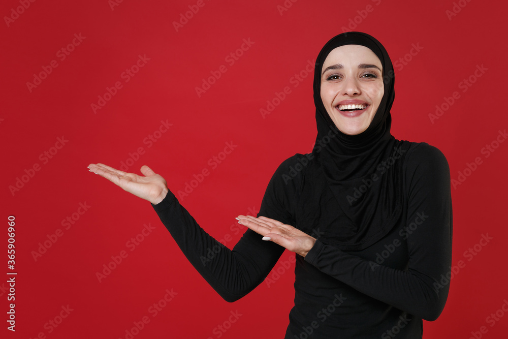Cheerful young arabian muslim woman in hijab black clothes posing isolated on bright red wall background studio portrait. People religious lifestyle concept. Mock up copy space. Pointing hands aside. Cheerful young arabian muslim woman in hijab black clothes posing isolated on bright red wall background studio portrait. People religious lifestyle concept. Mock up copy space. Pointing hands aside.
