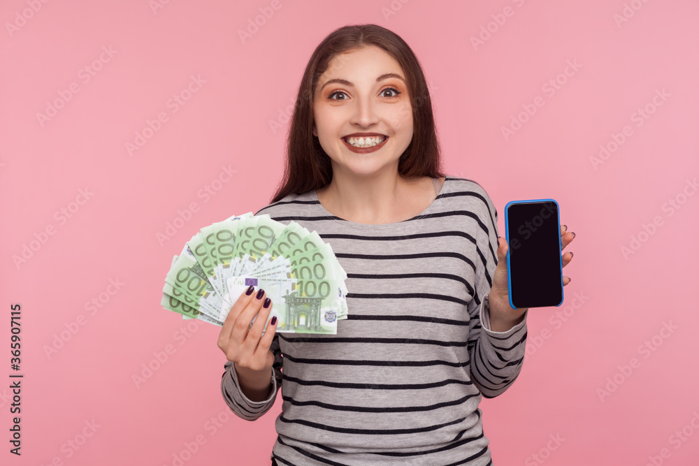 Mobile banking application. Portrait of happy woman in striped sweatshirt holding euro banknotes and cell phone with empty display, mock up for advertise. studio shot isolated on pink background Mobile banking application. Portrait of happy woman in striped sweatshirt holding euro banknotes and cell phone with empty display, mock up for advertise. studio shot isolated on pink background