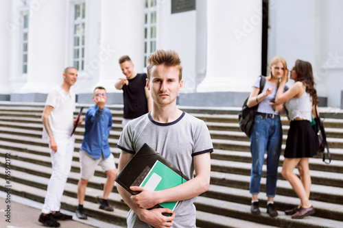 Fototapeta Naklejka Na Ścianę i Meble -  Young sad boy stand near university building and look into the camera, while other students behind show fingers at him, whisper and laugh. Education, bullying, social relations and people concept.