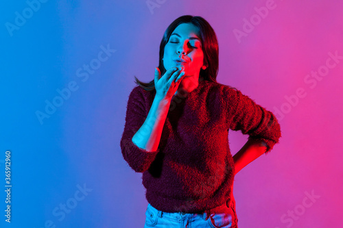 Neon light romantic portrait of lovely beautiful brunette woman standing with closed eyes, sending sensual air kiss, dreaming of love relations, affection feelings. indoor studio shot isolated
